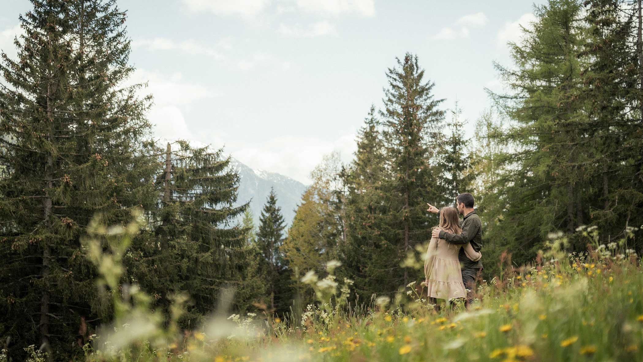 Ein Paar umarmt sich auf einer Wildblumenwiese, umgeben von hohen Kiefern und fernen Bergen. - Apartments & Chalet Selm