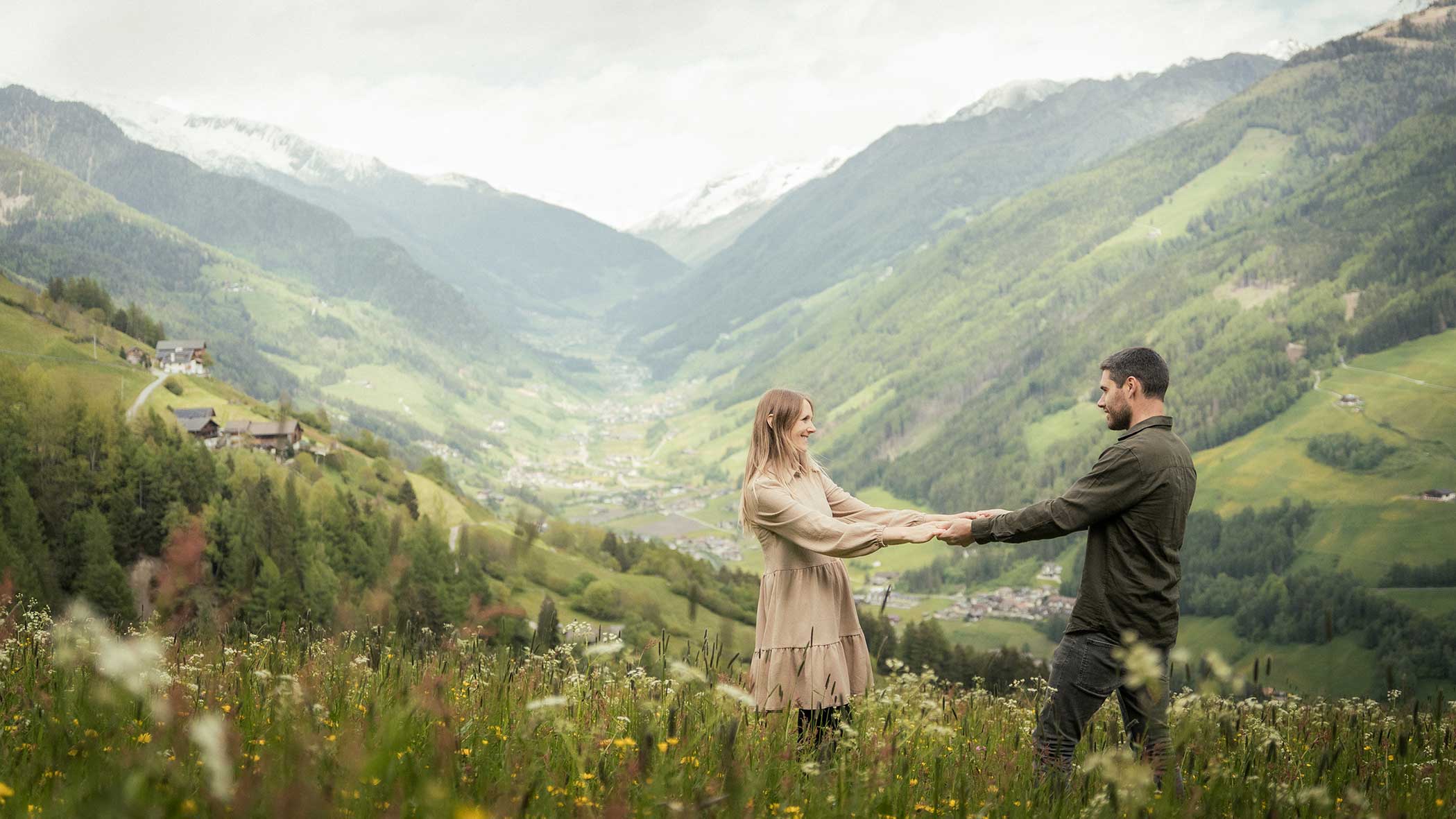 Ein händchenhaltendes Paar auf einer Blumenwiese mit Bergen und einem Tal im Hintergrund. - Apartments & Chalet Selm