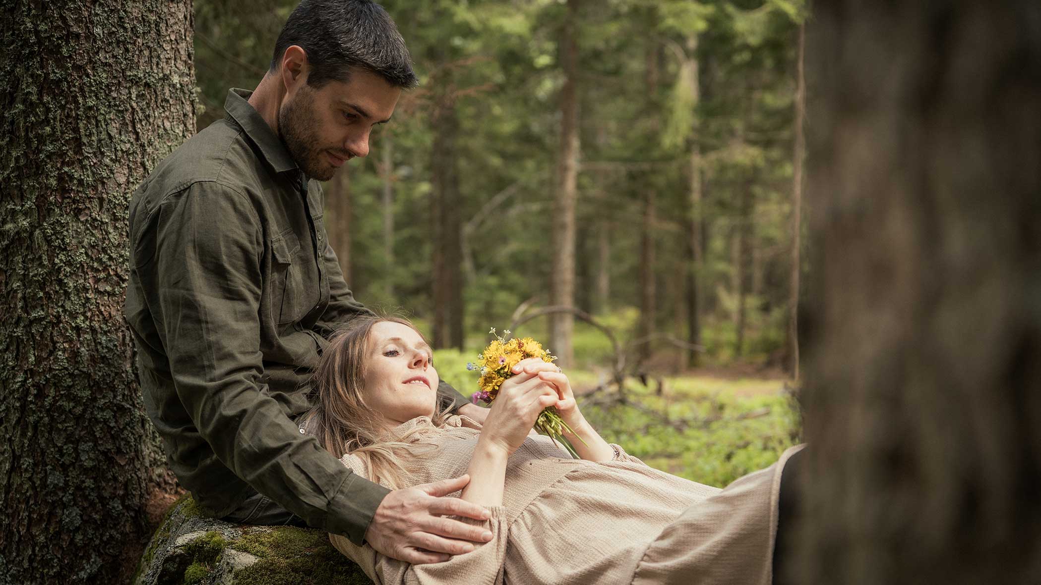 Ein Mann sitzt an einem Baum im Wald und hält eine Frau, die in seinem Schoß liegt und gelbe Blumen hält. - Apartments & Chalet Selm