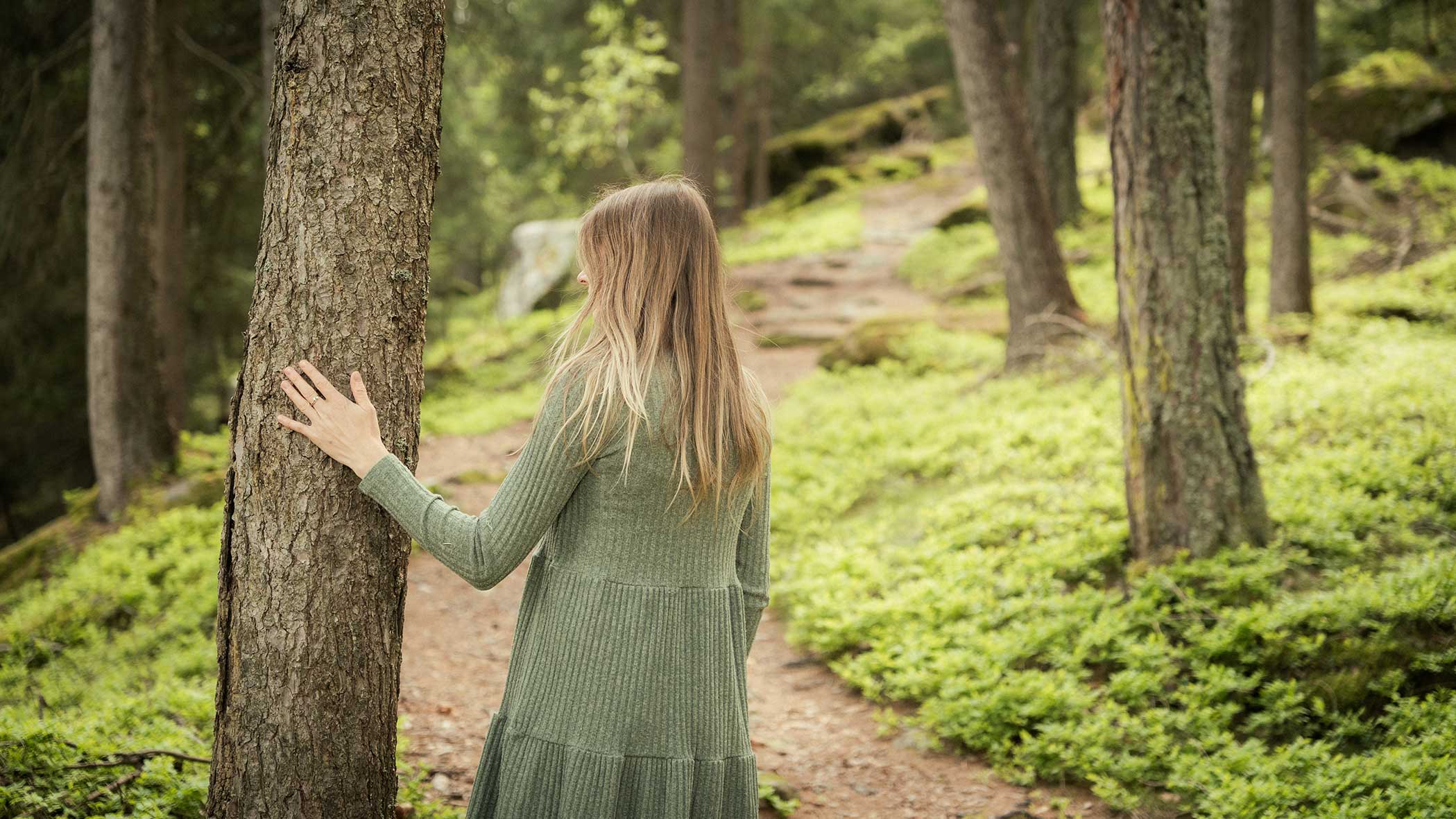 Frau in einem grünen Kleid, die einen Baum berührt, während sie auf einem Waldweg im Grünen spazieren geht. - Apartments & Chalet Selm