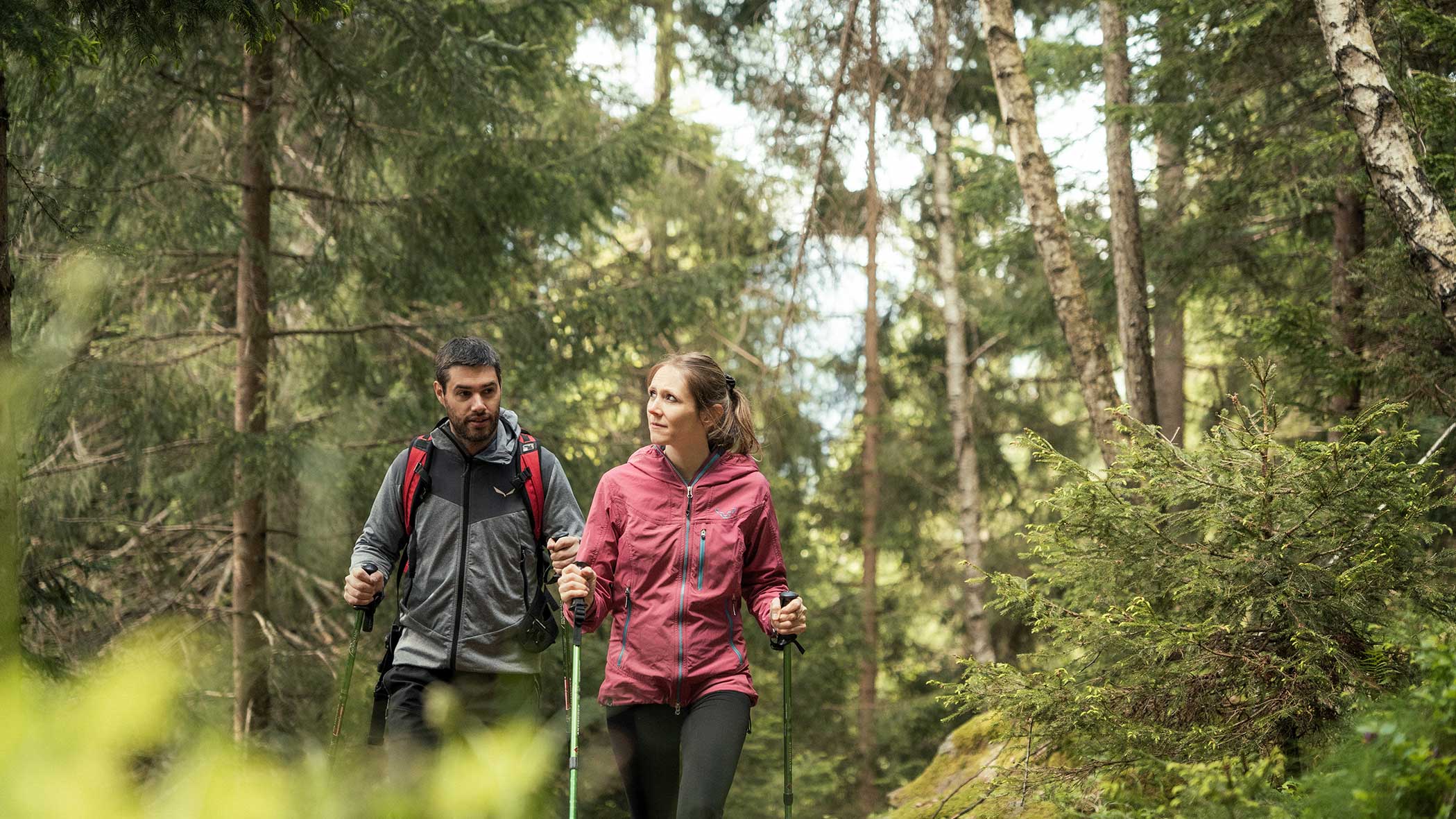 Zwei Personen wandern mit Trekkingstöcken durch einen Wald, umgeben von hohen Bäumen und viel Grün. - Apartments & Chalet Selm
