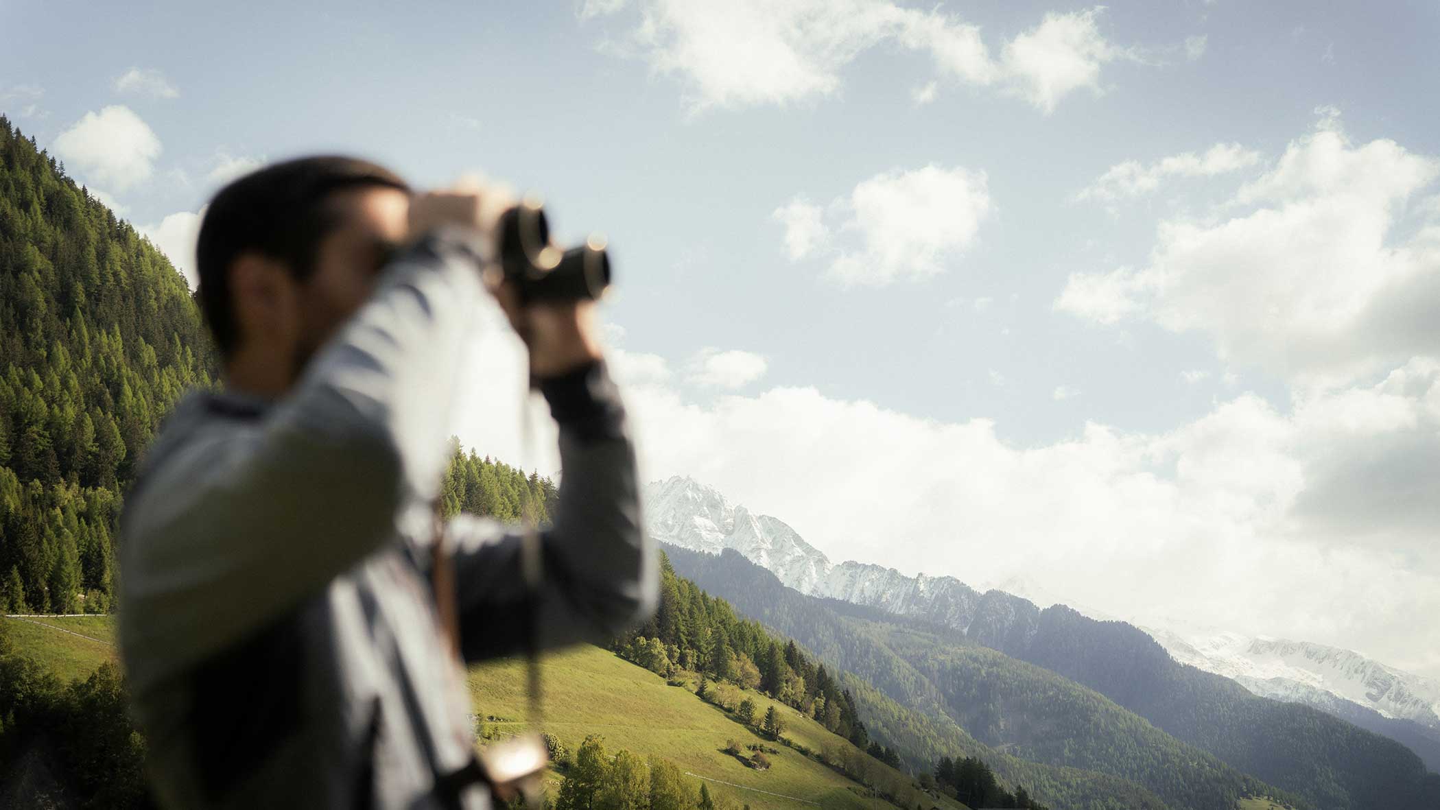Eine Person, die ein Fernglas benutzt, blickt auf Berge und grüne Hügel unter einem teilweise bewölkten Himmel. - Apartments & Chalet Selm