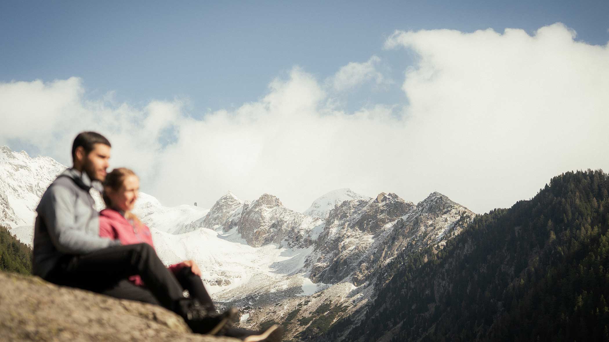Ein Paar sitzt auf einem Felsen und blickt auf schneebedeckte Berge und einen Wald unter einem teilweise bewölkten Himmel. - Apartments & Chalet Selm