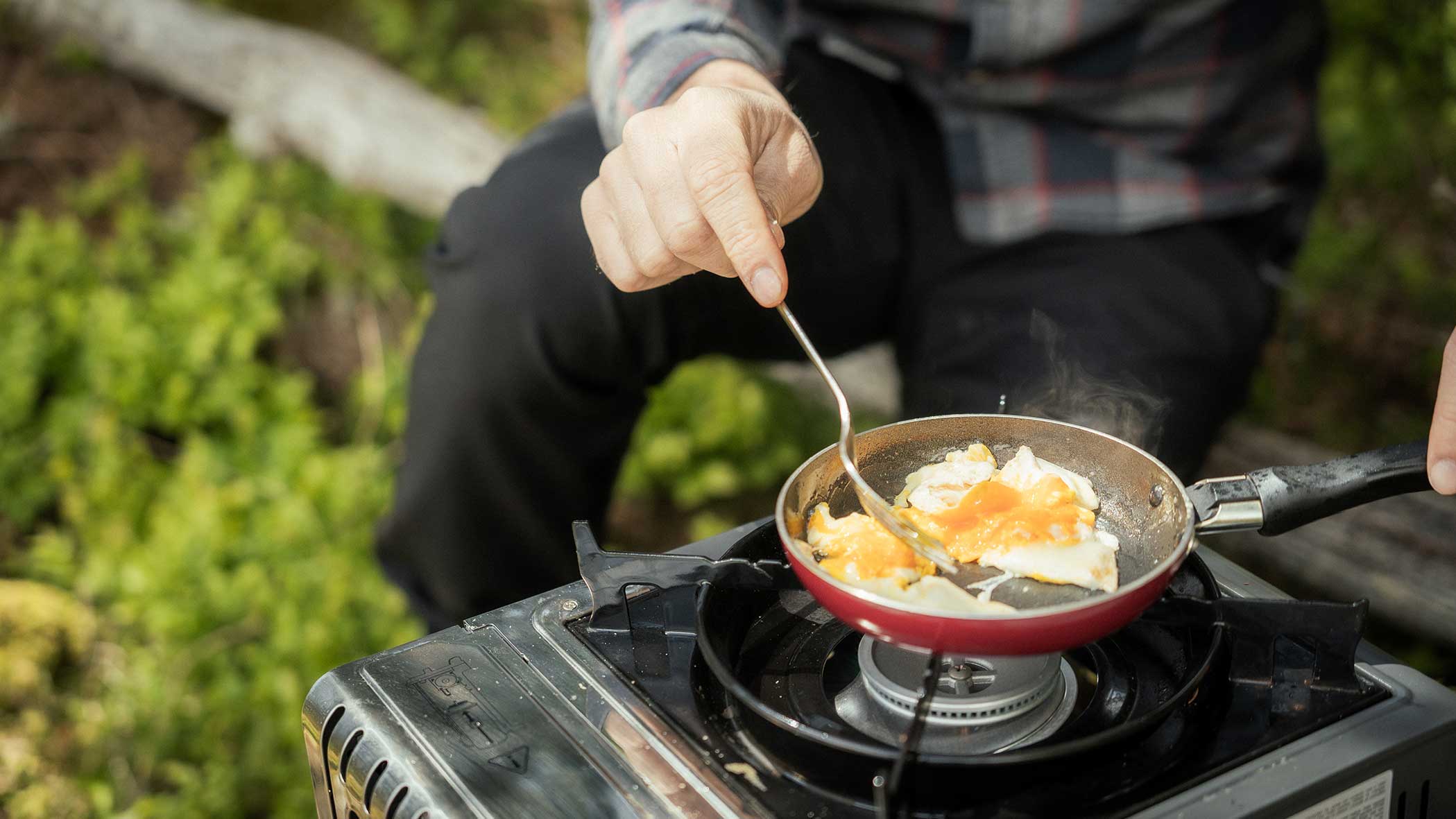 Eine Person kocht Eier in einer kleinen Pfanne auf einem tragbaren Kocher im Freien, umgeben von Grünpflanzen. - Apartments & Chalet Selm