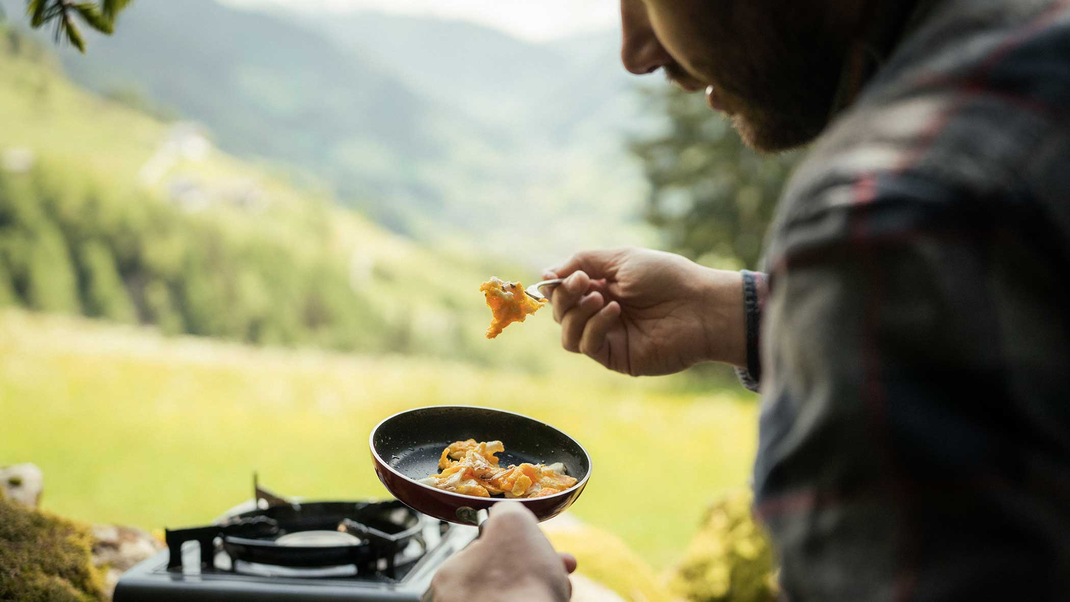 Eine Person, die im Freien in einer kleinen Pfanne Essen zubereitet, mit Bergen und Grün im Hintergrund. - Apartments & Chalet Selm