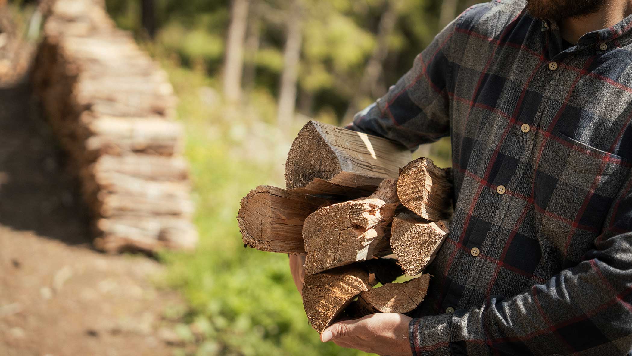 Mann im karierten Hemd trägt Brennholz, mit gestapelten Holzscheiten und Grünzeug im Hintergrund. - Apartments & Chalet Selm