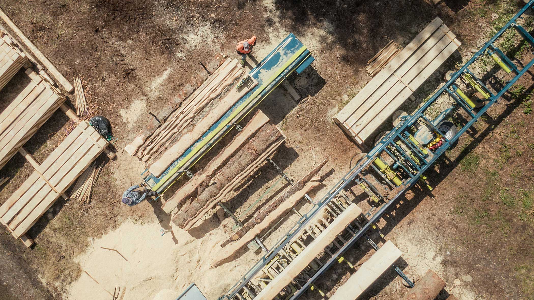 Luftaufnahme von zwei Personen, die in einem Sägewerk arbeiten, wobei Stämme und geschnittenes Holz auf dem Boden liegen. - Apartments & Chalet Selm