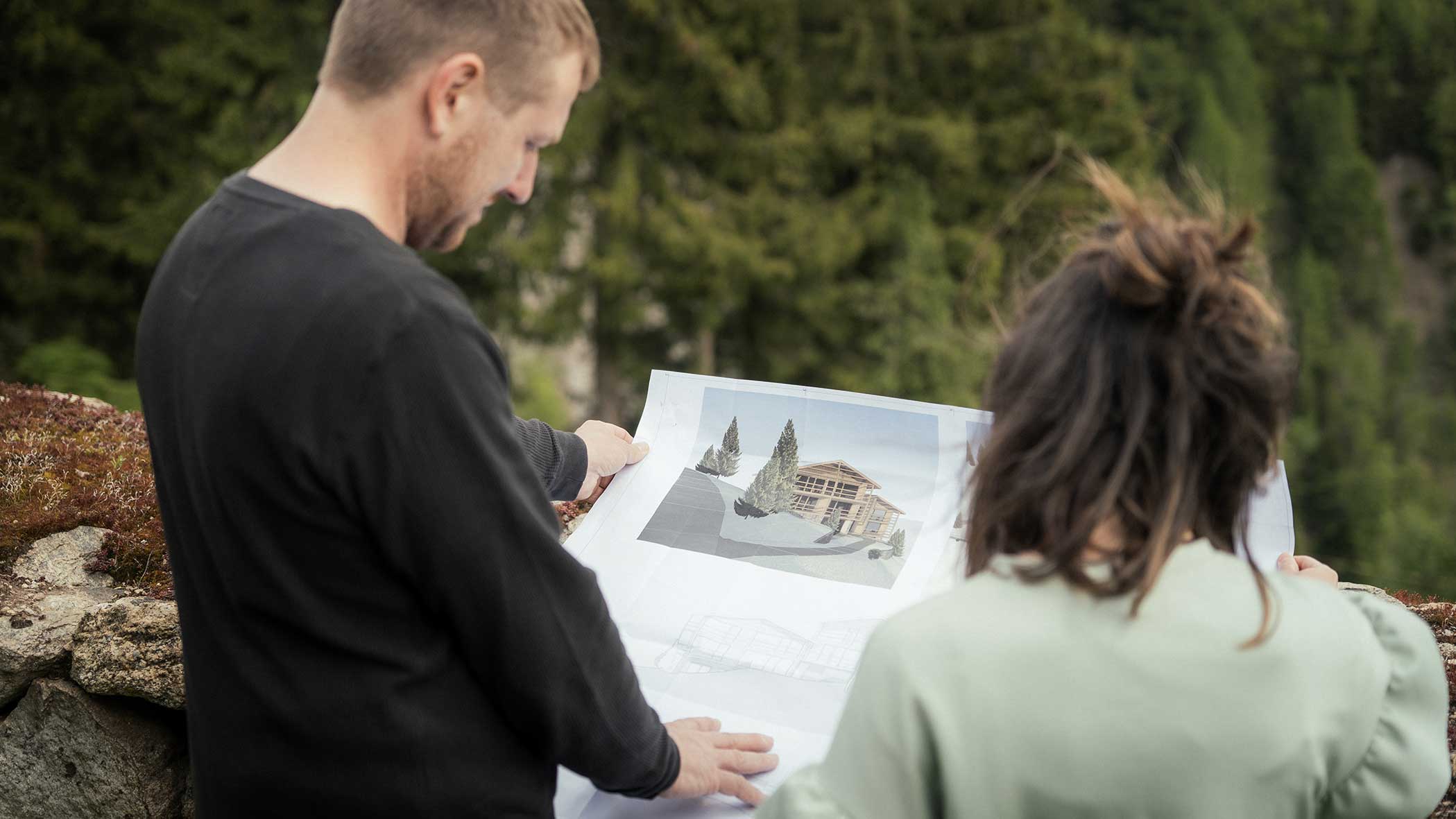 Zwei Personen im Freien, die sich Architekturpläne mit einem Gebäuderendering ansehen, umgeben von Bäumen. - Apartments & Chalet Selm