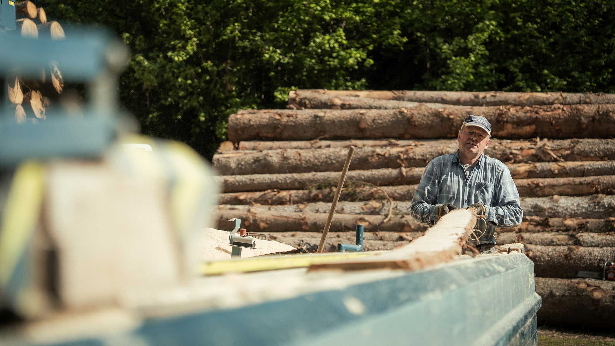 Ein Mann mit Mütze arbeitet mit Holzstämmen auf einem Holzlagerplatz, mit Stapeln von geschlagenem Holz im Hintergrund. - Apartments & Chalet Selm
