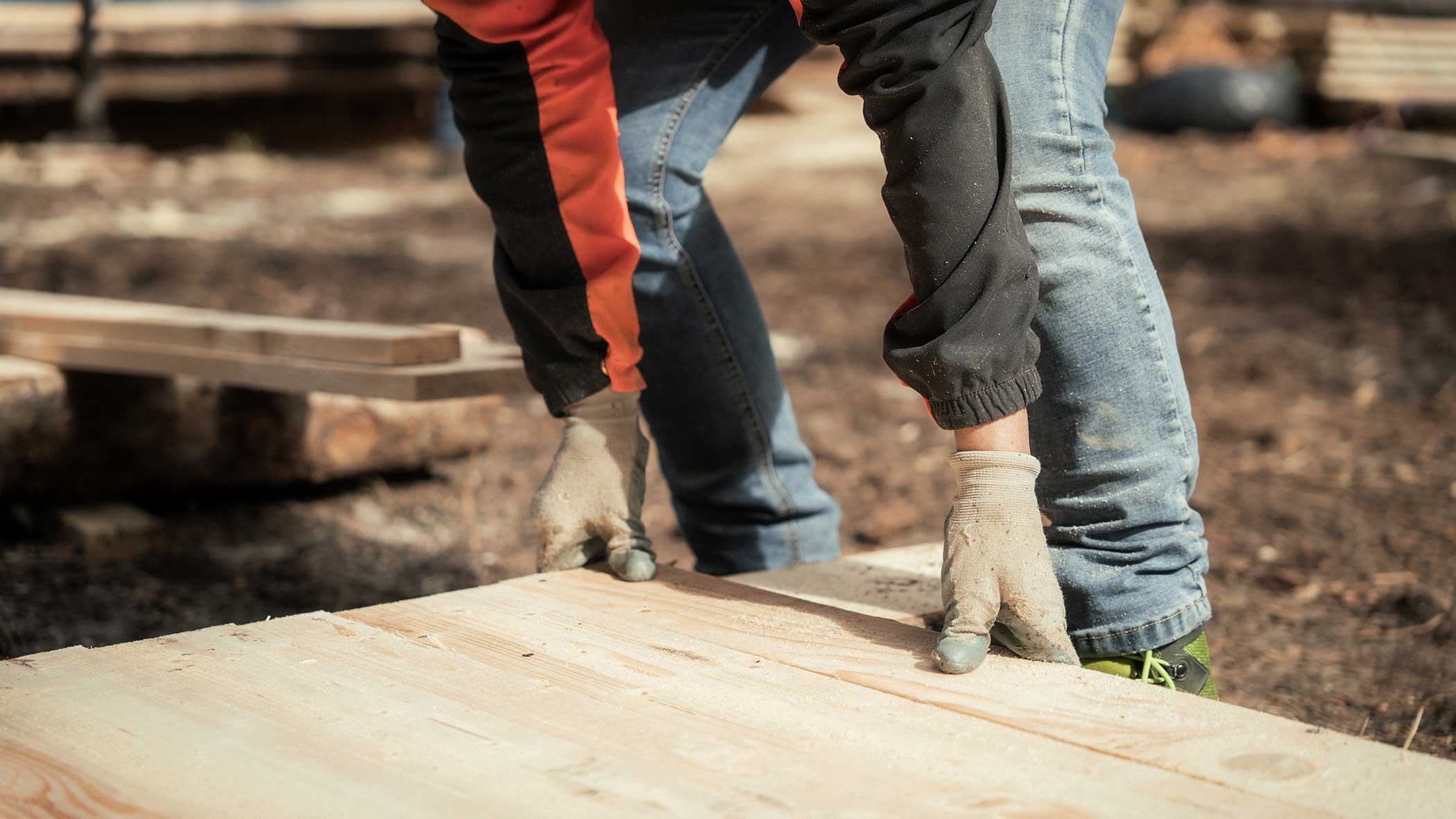 Eine Person mit Handschuhen hebt auf einer Baustelle im Freien eine Holzbohle an. - Apartments & Chalet Selm