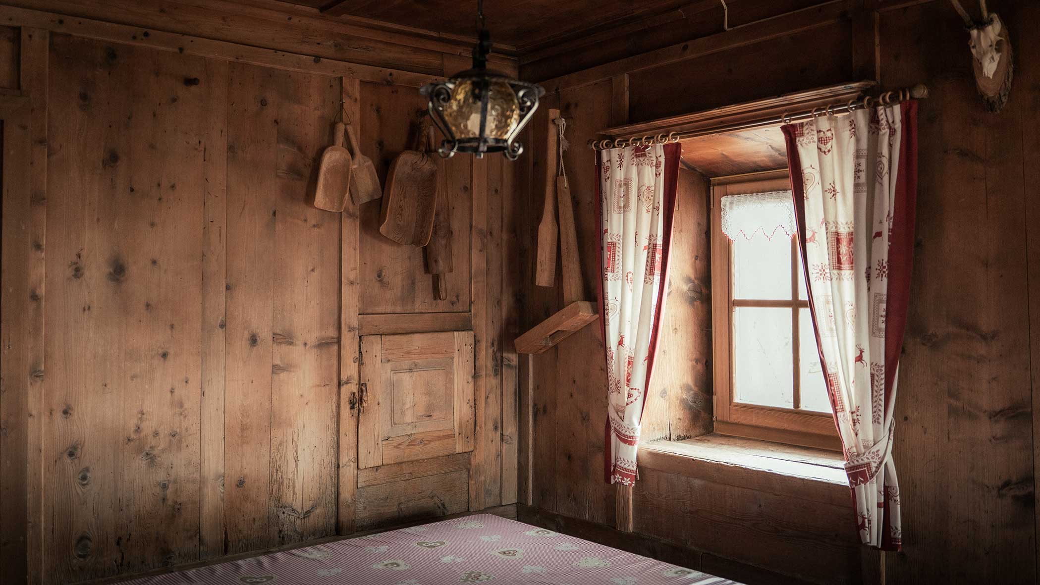 Ein rustikales Holzzimmer mit einem Fenster, gemusterten Vorhängen, hängenden Utensilien und einer Deckenlampe. - Apartments & Chalet Selm