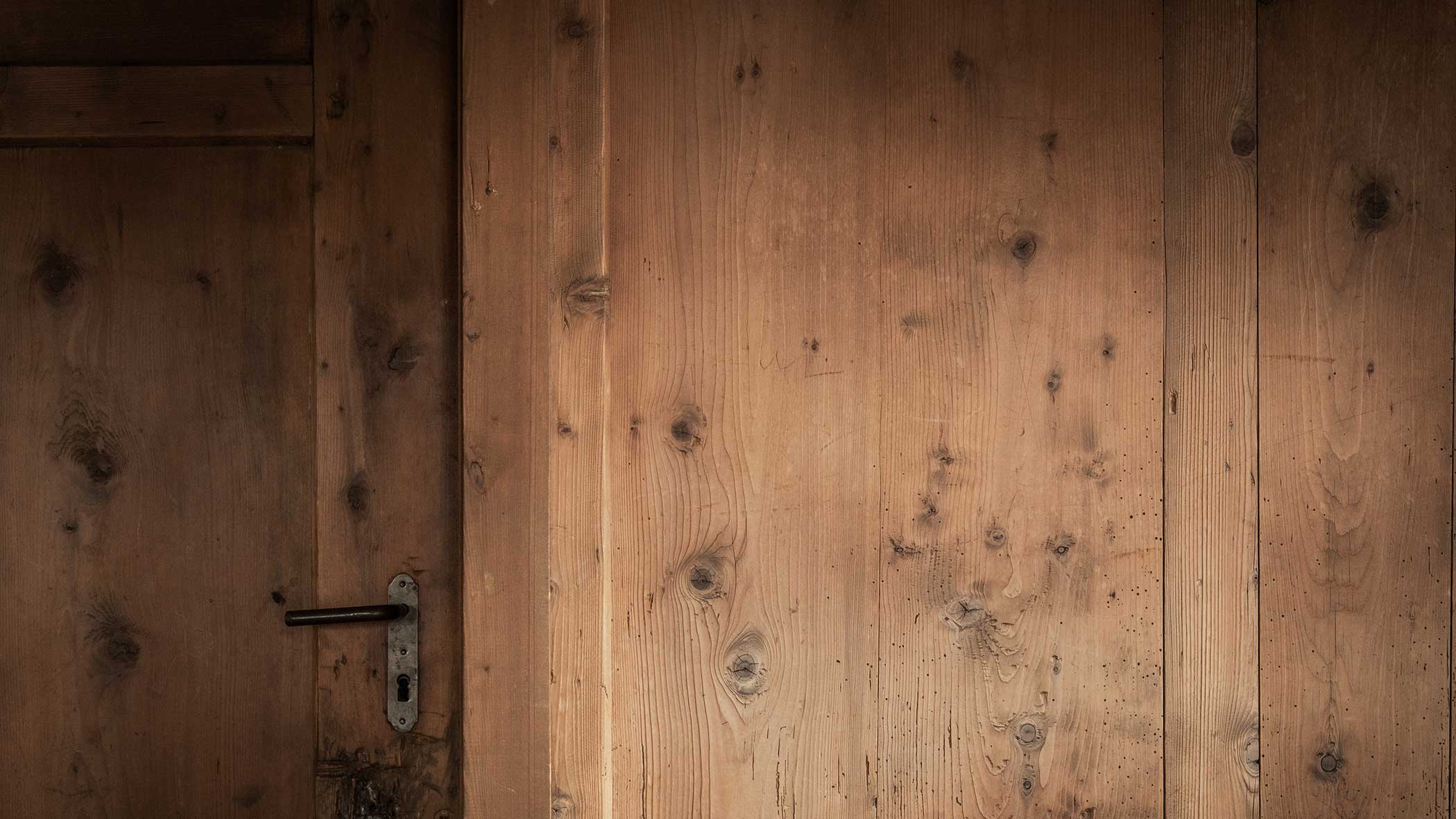 Nahaufnahme einer Holztür mit Metallgriff und eines Abschnitts einer holzgetäfelten Wand, der die natürliche Maserung zeigt. - Apartments & Chalet Selm